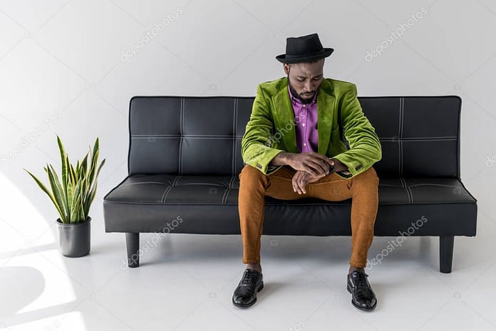 african american fashionable man in hat checking time while sitting on black sofa